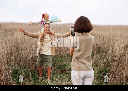 Mama macht Fotos von glücklichen Vater und Tochter im Sommer Natur auf dem Smartphone. Familienfreizeitreise mit Kleinkind auf beigefarbener Wiese Stockfoto