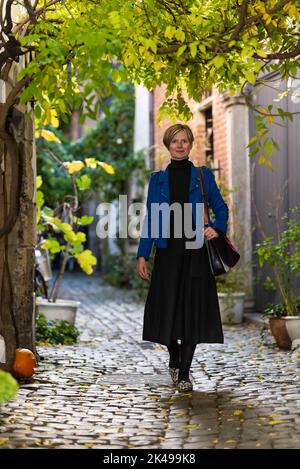 Attraktive 30-jährige weiße Frau in einem langen Kleid, die eine enge Straße mit Kopfsteinpflaster entlang geht, Brüssel Stockfoto