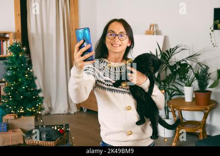 Junge Frau umarmt schwarze Hauskatze und macht Selfie in festlichen weihnachtslichtern in modernen Raum mit Geschenken und weihnachtsdekoration. Frohes Wint Stockfoto