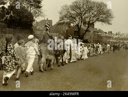 Jugendliche in ausgefallenen Kleidern, darunter ein Junge auf einem Esel, gehen am 6. Mai 1935, dem Silbernen Jubilee-Tag von König George V, in einer langen geraden Straße (wahrscheinlich Wakefield Road) entlang, die mit Fahnen geschmückt ist und sich in Denby Dale in West Yorkshire, England, Großbritannien, befindet. Stockfoto