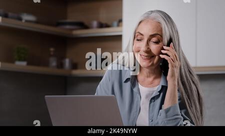 Weibliche leitende Geschäftsfrau mittleren Alters, grauhaarige Spezialistin, sitzt in der Küche und verhandelt per Telefon mit einem Geschäftspartner Stockfoto