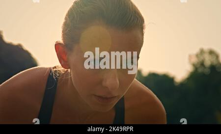 Motivierte junge Läuferin Weiße Jogginghose sitzt in gebeugter Position an der Startlinie und ist bereit, den Rennstrecken-Marathon zu joggen und sich auf den Sprint-Lauf vorzubereiten Stockfoto