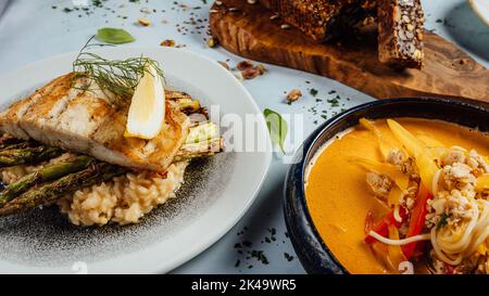 Eine Kürbiscremesuppe und gegrillter Lachs neben Nüssen und Samenbrot auf einem Tisch Stockfoto