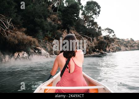 Junge kaukasische Frau von hinten sitzt in einem Kajak, paddelt herum, um die Bäume am Seeufer, tarawera See, Neuseeland zu erreichen Stockfoto