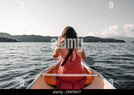 kaukasisches Mädchen von hinten in einem rosa Kleid sitzt in einem Kajak paddeln auf dem See in Richtung der Berge und Wald, tarawera See, neu Stockfoto