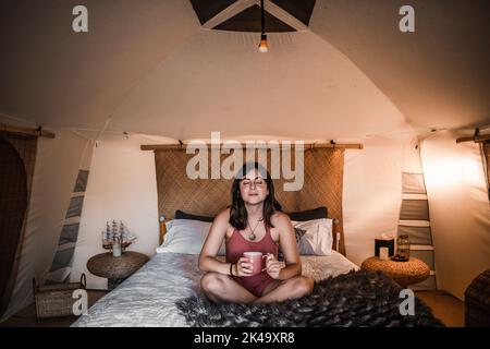 Entspannte, glückliche, ruhige kaukasische junge Frau, die im Zelt auf dem Bett mit gekreuzten Beinen eine Tasse Kaffee in den Händen hält Stockfoto