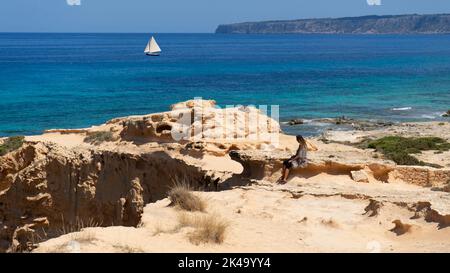 Panoramablick auf eine Frau, die auf Felsen sitzt, während sie ein Segelschiff auf dem Meer beobachtet. Insel Formentera, Spanien Stockfoto