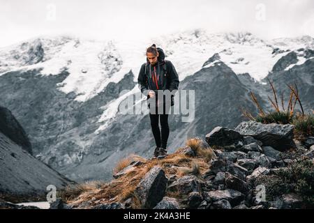 Junger Kaukasier warm in einer Jacke bequeme Turnschuhe und schmale schwarze Hosen mit einer hängenden Kamera, die den Boden in der Nähe von Schnee vorsichtig anschaut Stockfoto