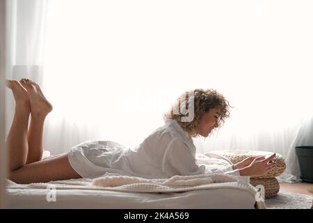 Fröhliche junge Dame, die lächelt und im Bett auf dem Handy stöbert Stockfoto