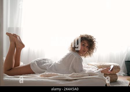 Selbstbewusste junge barfüßige Frau, die sich morgens mit dem Smartphone auf dem Bett entspannt Stockfoto