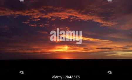 Wolken, die Sonnenlicht reflektieren, nachdem die Sonne unter den Horizont untergegangen ist, in rosa und orangen Farbtönen über der silhouettierten, dunklen Erde Stockfoto