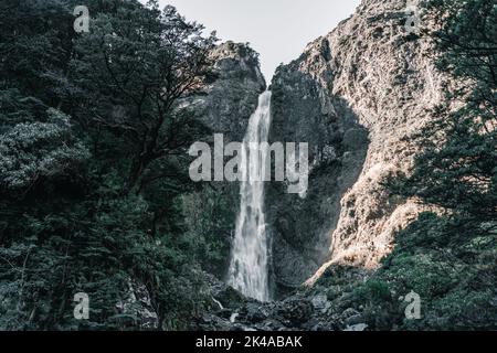 Hoher, enger Wasserfall mit wenig Wasser zwischen den Felsen der Berge, der von Sonnenlicht unter einem grauen Sonnenuntergangshimmel in der Nähe der Waldbäume beleuchtet wird Stockfoto