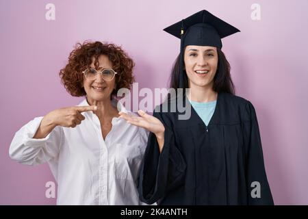 Hispanische Mutter und Tochter mit Abschlusskappe und Zeremoniengewand staunten und lächelten der Kamera zu, während sie mit der Hand vorstellten und mit Fi zeigten Stockfoto