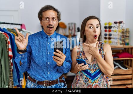 Interrassisches Paar im mittleren Alter im Einzelhandel mit Smartphone überrascht, mit dem Finger zur Seite zeigend, offener Mund erstaunt Ausdruck. Stockfoto