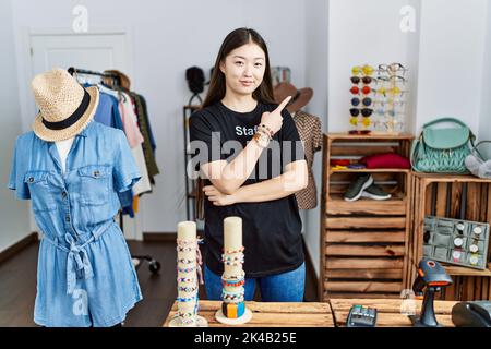 Junge asiatische Frau, die als Managerin in der Einzelhandelsboutique arbeitet und mit dem Finger zur Seite zeigt, zeigt Werbung, ernstes und ruhiges Gesicht Stockfoto
