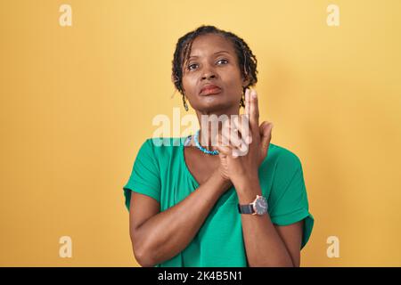Afrikanische Frau mit Dreadlocks, die auf gelbem Hintergrund stehen und symbolische Waffe mit Handbewegung halten, Waffen töten, wütendes Gesicht spielen Stockfoto