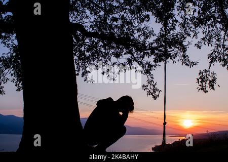 Baum und Silhouette des Menschen. Der Mann, der unter dem Baum hockend sitzt. Deprimierter Mann. Stockfoto