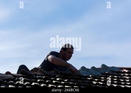 Mann repariert Dachziegel mit Zement auf einem alten Gebäudedach im Dorf an einem sonnigen Tag. Silhouette eines Mannes auf einem Dach während der Arbeit Stockfoto