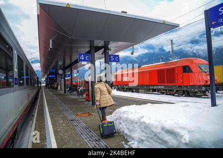 02-21-2022 Salzburg, Österreich. Die Plattform irgendeines Bahnhofs . Passagier mit Koffer auf RadenZug links und Güterzug rechts- rot elektr Stockfoto