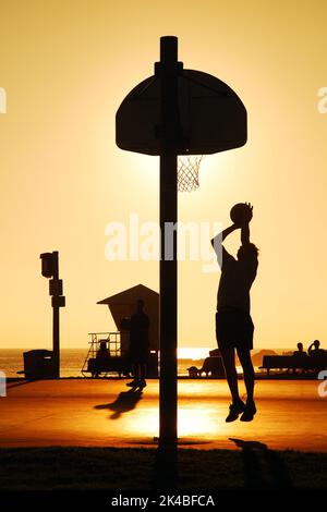 Ein erwachsener Mann schieß auf einen Basketballplatz im Freien, während die Sonne untergeht Stockfoto