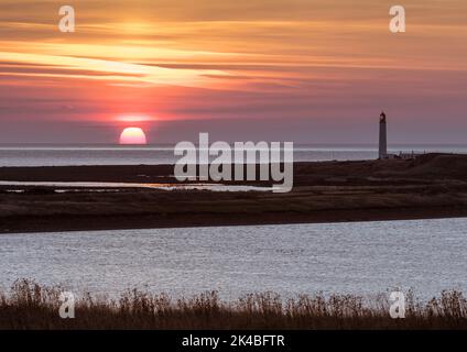 Goldener Sonnenorb, der teilweise am Meereshorizont aufging, vom Barns Ness Point aus gesehen, mit Leuchtturm und Küstenlagune im Vordergrund. Stockfoto