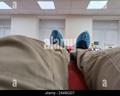 Die Beine sind in Stiefeln und blauen Überziehschuhen verkrackt. Überschuhe an den Beinen aus nächster Nähe. Hygienische Standards im Krankenhaus. Mann in chirurgischen Stiefeln in Spende Stati Stockfoto