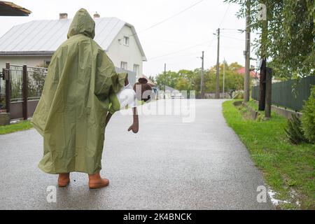 Junge mit grünem Regenmantel, der an regnerischen Tagen auf der Straße steht. Kinderportrait im Herbst kaltes Wetter. Hält weiches Affen Spielzeug. Platz für Text kopieren. Stockfoto