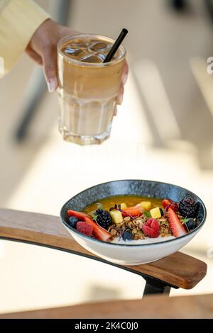 Eine vertikale Aufnahme einer Smoothie-Schüssel mit Beeren und einem Glas Eiskaffee in der Hand einer Frau Stockfoto