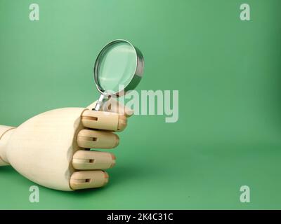 Eine hölzerne Hand, die eine Lupe auf einem grünen Hintergrund hält Stockfoto