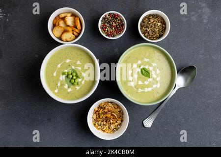 Horizontales Bild von zwei Schalen mit Erbsensuppe und Brokkoli, mit Toast, Samen und Pfefferkörnern auf Schiefer Stockfoto