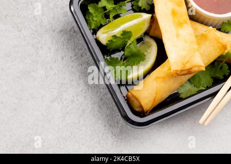 Blick von oben auf asiatische Frühlingsrollen, geschnittene Limette, Chilisauce und Essstäbchen auf grauem Hintergrund Stockfoto