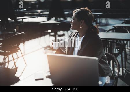 Eine Profilansicht einer hinterleuchteten Silhouette einer reifen, schönen hispanischen Unternehmerin, die mit ihrem Laptop in einem Straßencafé sitzt, während sie ihren Cof hat Stockfoto