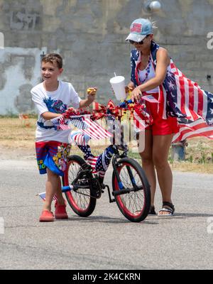 Eau Claire MI USA, 4 2022. Juli; Frau und kleiner Junge mit einem patriotischen Fahrrad geben Süßigkeiten aus, während sie in einer Parade vom 4.. Juli fahren Stockfoto