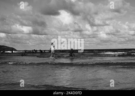 Eine Graustufenaufnahme von Menschen, die an der Strandküste unter einem bewölkten Himmel spielen Stockfoto