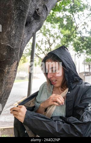 Junge Frau, die bei einem plötzlichen, unerwarteten Regen Regenmantel anlegt, in einem nassen Baum, mexiko, Schutz bietet Stockfoto