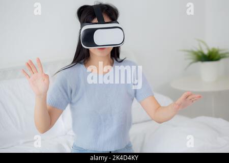 Junge asiatische Frau mit vr-Headset spielen Spiel mit aufgeregt auf dem Bett im Schlafzimmer zu Hause, weiblich mit Virtual Reality oder Metaverse Innovation für Stockfoto