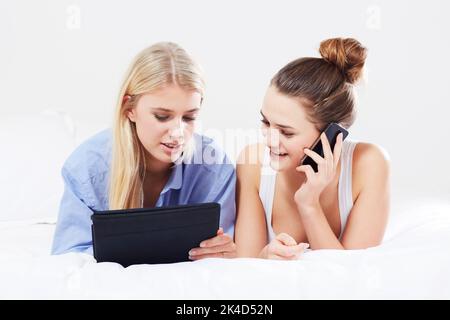 Genießen Sie die neuesten Gadgets. Eine schöne junge Frau, die auf ihrem Smartphone spricht, während ihre Freundin ein Tablet benutzt, während sie im Bett liegt. Stockfoto