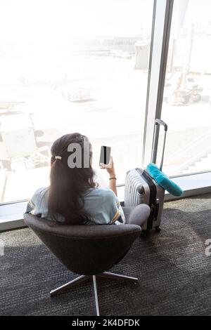 Die schwarzhaarige kaukasische Frau sitzt auf einem Sitz in der Lounge des Flughafens und überprüft ihr Handy. Sie hat sie auch weitergemacht. Langeweile. Stockfoto