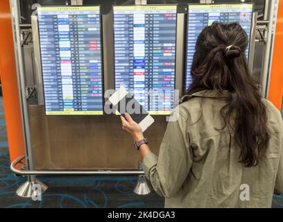 Die schwarzhaarige kaukasische Frau, die ihr Mobiltelefon in der Hand hält, sieht die Bildschirme für die an- und Abreise des Fluges am Flughafen an. Sie ist allein Stockfoto