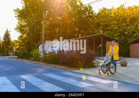 Junge Mutter, die mit ihrer Tochter den Rollstuhl schiebt, ein Mädchen, das mit einer zerebralen Lähmung lebt und auf dem Weg zur Therapie die Straße überquert. Stockfoto