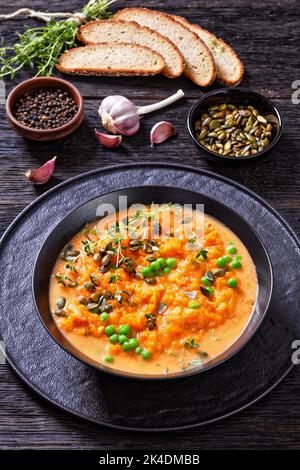 Nahaufnahme einer dicken Süsskartoffelsuppe mit Gemüse in einer schwarzen Schüssel auf einem dunklen Holztisch mit Kräutern, Pfefferkörnern, Kürbiskernen und Roggenbrotscheibe Stockfoto