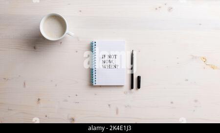 Draufsicht auf einen spiralförmigen Notizblock mit einem „If Not Now When“-Zeichen. Schwarzer Marker und eine Tasse Kaffee auf einem Schreibtisch daneben. Stockfoto