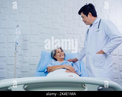 Glückliches Lächeln Asiatische ältere, ältere Patientin in hellblauem Kleid, die mit Kochsalzlösung auf dem Bett lag, während der Arzt in weiß ihr Sho berührte Stockfoto