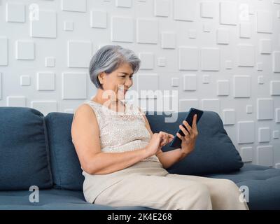 Glückliche asiatische ältere Frau sitzt auf der Couch, hält und schaut auf den Handybildschirm in der Hand im Wohnzimmer. Ältere Frauen, die ein Smart-Gerät, einen Computer, benutzen Stockfoto