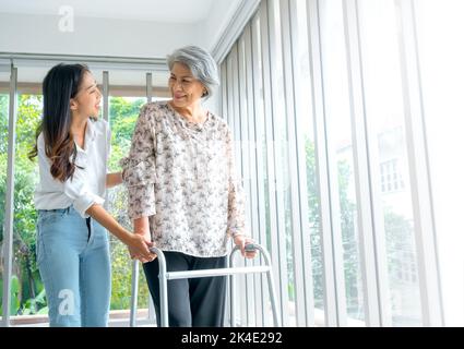 Pfleger, lächelnd junge asiatische Frau, Tochter oder Enkelkind Unterstützung helfen ältere Frau, Mutter oder Großeltern versuchen, mit Walker Rahmen am h gehen Stockfoto