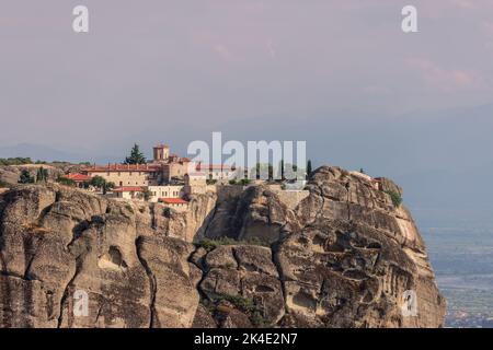 Das Holy Trinity Monastery krönt einen verwitterten Felspfeiler in Meteora, Griechenland, mit roten Ziegeldächern, einer Kirchkuppel und Zypressen über einem nebligen Tal. Stockfoto