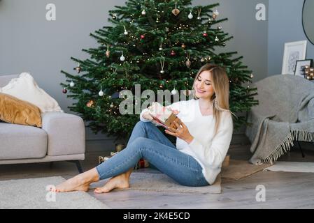 Die reizende Frau sitzt auf dem Boden in der Nähe des Weihnachtsbaums im Wohnzimmer und hält ein Überraschungsgeschenk in ihren Händen Stockfoto