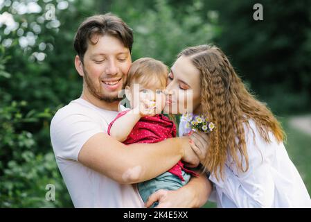 Glückliche junge schöne Familie mit einem jungen Sohn genießt einen wunderschönen Tag in einem Sommerpark. Junge Eltern küssen ein Kind Stockfoto