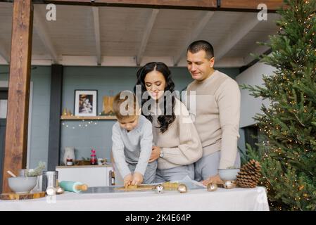 Mama, Papa und kleiner Sohn schneiden Formen aus Teig für Ingwerkekse oder Lebkuchenhaus aus. Vorbereitung auf Weihnachten Stockfoto