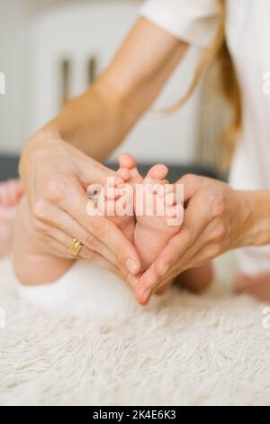 Die liebevolle Mutter hält in ihren fürsorglichen Händen die Beine und Finger eines kleinen Neugeborenen, eines schlafenden Kindes in Nahaufnahme auf dem Bett. Selektiver Fokus. Frauenhap Stockfoto